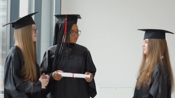 Три студентки різних рас з дипломом в руках. Випускники чорного одягу та квадратних капелюхів розмовляють. Концепція освіти — стокове відео