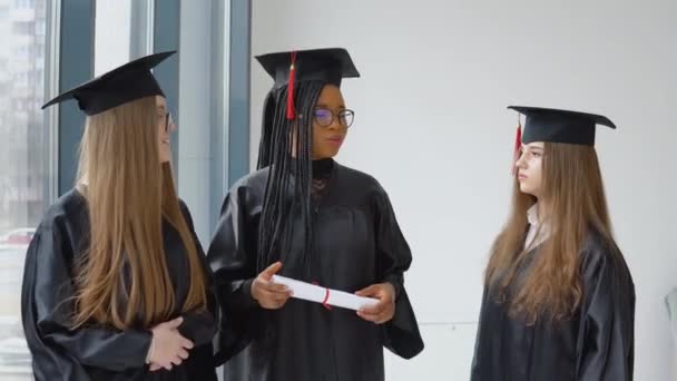 Trzy studentki rasy kaukaskiej i afrykańskiej z dyplomem w rękach. Absolwenci w czarnych szatach i kwadratowych kapeluszach komunikują się ze sobą. Koncepcja edukacji — Wideo stockowe