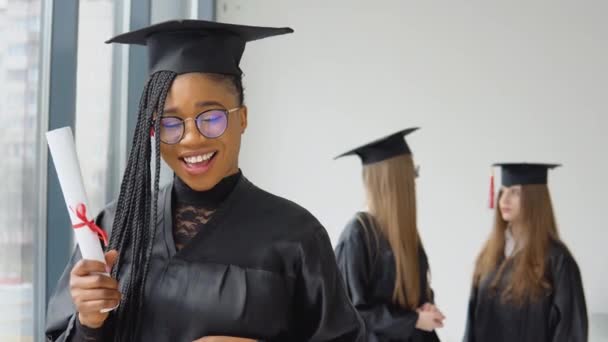 Um graduado alegre com um diploma na mão está na universidade contra o pano de fundo de graduados conversando uns com os outros. Programa de intercâmbio de estudantes entre universidades de todo o mundo — Vídeo de Stock