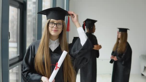 The graduate holds a diploma in one hand and a hat in the other. Masters degree in the university on the background of two female students of african american and caucasian races — Stock videók