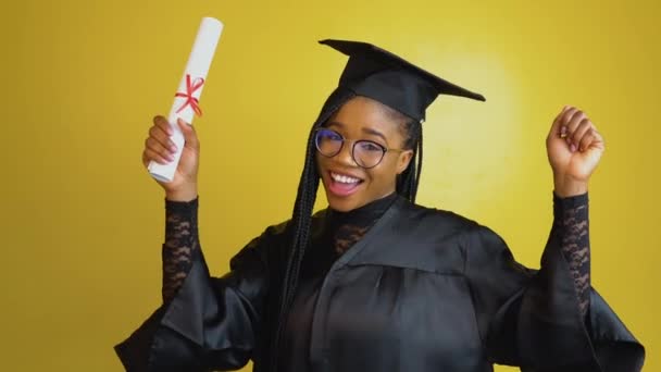 A joyful graduate student holds an international diploma and expresses happiness and positive emotions. Student in a black gown and hat on a yellow solid background. Education concept — Stock videók