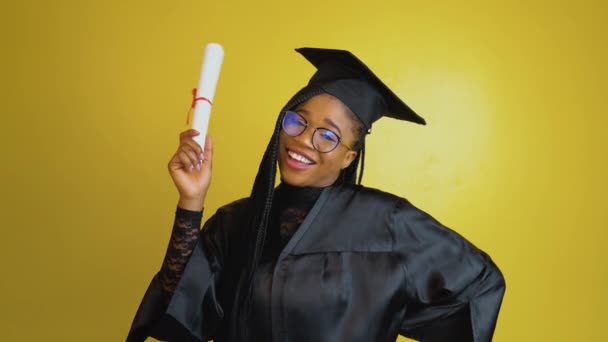 Радостный аспирант имеет международный диплом и выражает счастье и позитивные эмоции. Студент в черном халате и шляпе на жёлтом фоне смотрит в камеру. Болонский процесс — стоковое видео