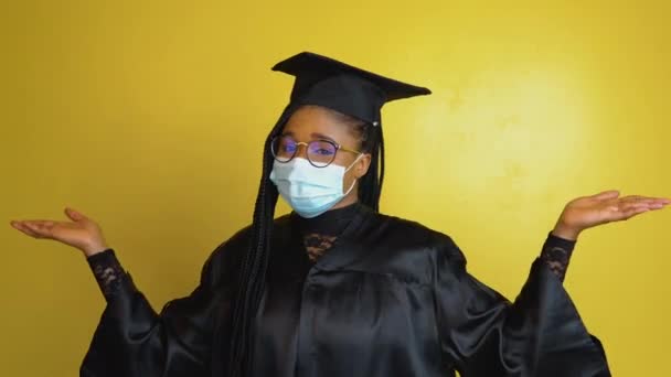 Den studerende i en beskyttende maske udtrykker med fagter behovet for at overholde karantæne sikkerhedsforanstaltninger. Studerende i en sort kåbe og hat på en gul solid baggrund ser på kameraet – Stock-video