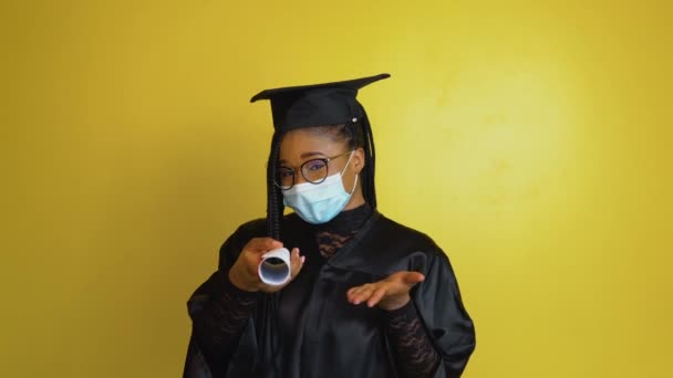 Un estudiante graduado en una máscara protectora apunta con un diploma en la mano hacia la cámara. Estudiante con una bata negra y sombrero sobre un fondo sólido amarillo mira a la cámara — Vídeos de Stock