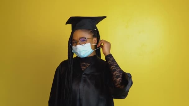 African american female student in a black robe and hat on a yellow solid background looks at the camera and removes the protective mask from his face — Stock videók