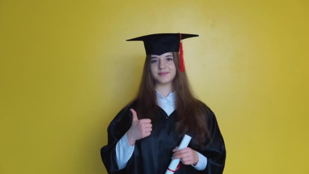 Caucásico chica estudiante muestra el dedo hacia arriba mientras se queda delante de la cámara con graduarse diploma en sus manos — Vídeo de stock