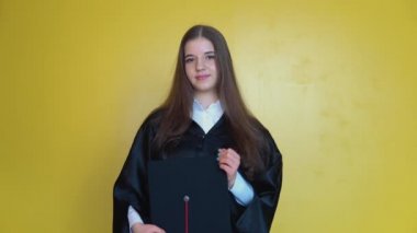 Bir üniversite mezunu kameranın önünde elinde kırmızı püsküllü bir usta şapkası ile duruyor. Sarı arka planda uzun düz saçlı genç bir kadının ön çekimi.