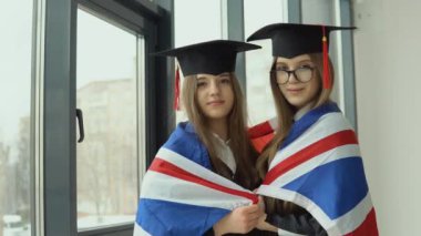 İki genç mutlu mezun kadın omuzlarında İngiliz bayrağı ile omuz omuza duruyorlar.