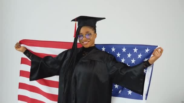 Estudante afro-americana elegante feliz em dança uniforme de pós-graduação segurando bandeira dos EUA sobre os ombros. Educação dos EUA — Vídeo de Stock