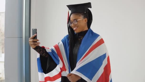 Feliz estudiante afro-americana con estilo en uniforme de graduado tomando selfie sosteniendo bandera británica sobre los hombros — Vídeo de stock