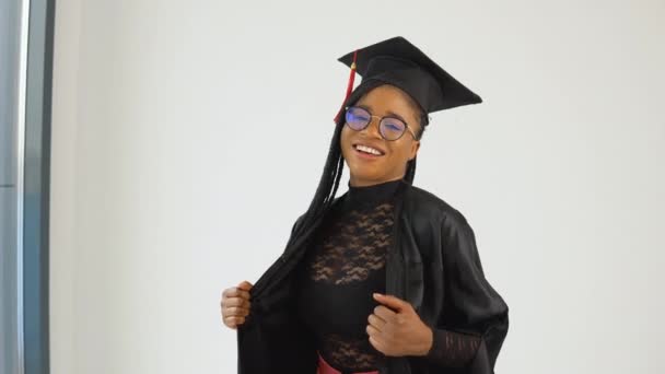 Feliz sexy mujer afroamericana elegante estudiante en uniforme de graduado de pie y coqueteando sobre un fondo blanco — Vídeo de stock