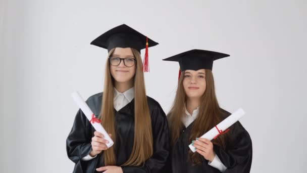 Dos jóvenes graduadas felices están hombro con hombro con diplomas de graduación en sus manos. Vista de cerca sobre fondo blanco — Vídeo de stock