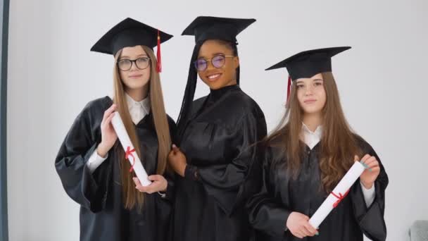 Три молодые счастливые выпускницы различных национальностей стоят бок о бок с дипломами в руках. Закрыть вид на белом фоне — стоковое видео