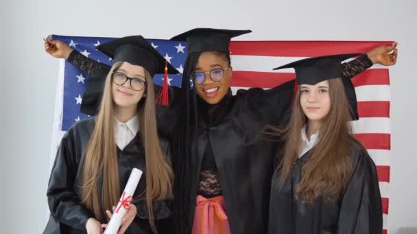 Drie vrouwelijke studenten in afgestudeerden pakken en hoeden houden de Amerikaanse vlag achter hun sholders. Studenten van verschillende rassen met de vlag van de VS — Stockvideo