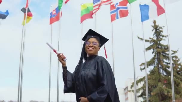Молодая афроамериканка радуется и меняет положение руки, в которой она имеет диплом о высшем образовании. Аспирант выступает против неба и флагов разных стран — стоковое видео