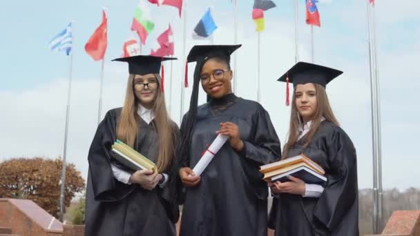 Três jovens universitárias graduadas de diferentes nacionalidades sorriem e se reúnem segurando livros e um diploma em suas mãos contra muitas bandeiras de diferentes países. — Vídeo de Stock