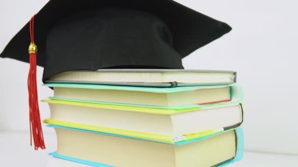 Un sombrero de graduado cuadrado negro con una borla roja se encuentra en una pila de libros. Los objetos giran alrededor de su eje en el soporte. Concepto educativo — Vídeo de stock