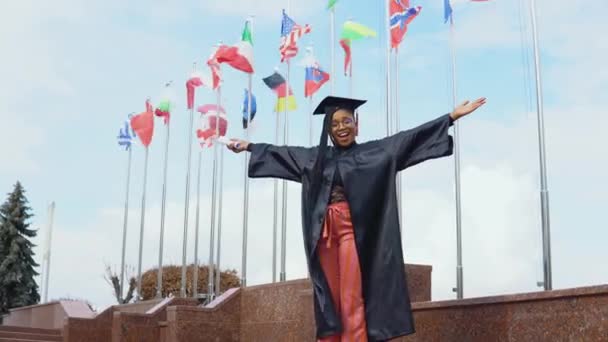 Genç Afrikalı Amerikalı kadın, Avrupa ve dünyadaki farklı ülkelerin bayraklarına karşı elinde yüksek öğrenim diplomasıyla seviniyor. Eczane Ustası — Stok video