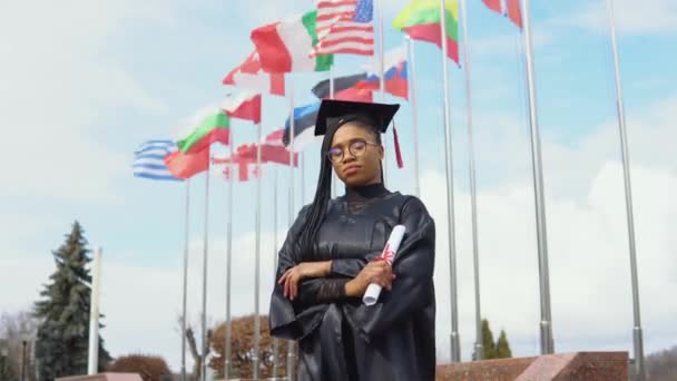 Jovem afro-americana se alegra com um diploma de ensino superior em sua mão em pé contra o céu e bandeiras de diferentes países agitando-se ao vento. Mestre das Leis — Vídeo de Stock