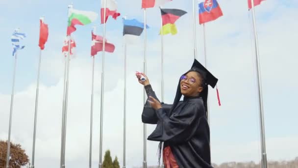 Una joven afroamericana se regocija y señala con un diploma a las banderas de diferentes países ondeando al viento — Vídeo de stock