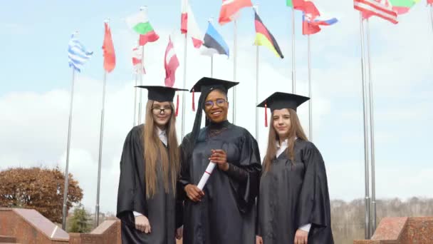 Três jovens mulheres de diferentes nacionalidades sorriem contra muitas bandeiras de diferentes países agitando ao vento ao fundo. Vista ao ar livre — Vídeo de Stock