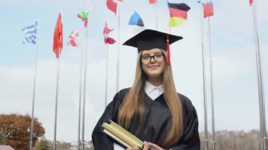 Bir üniversite mezunu, elinde kitaplarla dünyanın bayraklarının arkasında duruyor. Açık alanın görüntüsü. Mavi gökyüzünde ulusal bayraklar