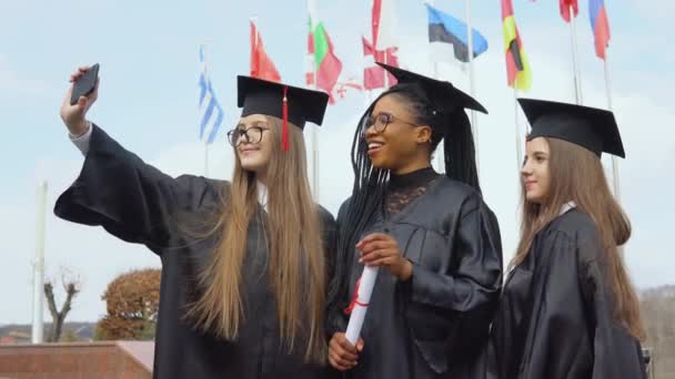 Drei junge Frauen verschiedener Nationalitäten lächeln und machen Selfies vor vielen Flaggen verschiedener Länder, die im Hintergrund im Wind flattern. Blick ins Freie — Stockvideo