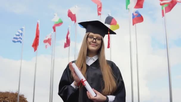 Bir üniversite mezunu, yüksek lisans diplomasını elinde tutan dünyanın bayraklarının arkasında duruyor. Açık alanın görüntüsü. Mavi gökyüzünde ulusal bayraklar — Stok video