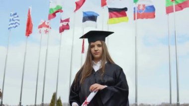 Bir üniversite mezunu, yüksek lisans diplomasını uluslararası standartlarda elinde tutan dünyanın bayraklarının arkasında duruyor. Açık alanın görüntüsü. Mavi gökyüzünde ulusal bayraklar