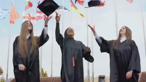 Farklı milletlerden üç genç kadın arka planda rüzgarda dalgalanan farklı ülkelerin bayraklarına neşe içinde şapkalar fırlatıyor. Açık hava görünümü — Stok video