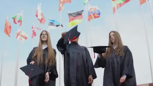 Tres mujeres jóvenes de diferentes nacionalidades alegremente arrojan sombreros contra muchas banderas de diferentes países ondeando al viento en el fondo. Vista exterior — Vídeos de Stock