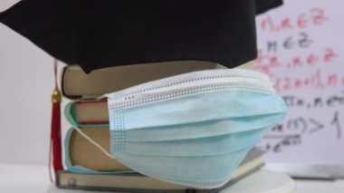 Efendinin siyah kare şapkası bir sütuna dizilmiş ve koruyucu bir tıbbi maskeyle süslenmiş ders kitaplarının üzerinde yatıyor. Üniversite mezununun nitelikleri duruşunda kendi ekseni etrafında döner.