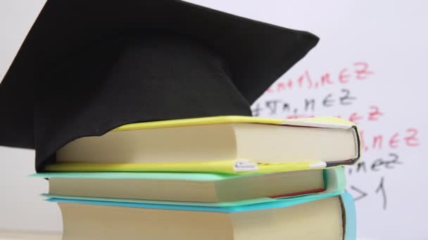 De zwarte vierkante hoed van de meester ligt op de leerboeken gestapeld in een kolom. De attributen van een universitair afgestudeerde draaien om zijn as op een standaard — Stockvideo