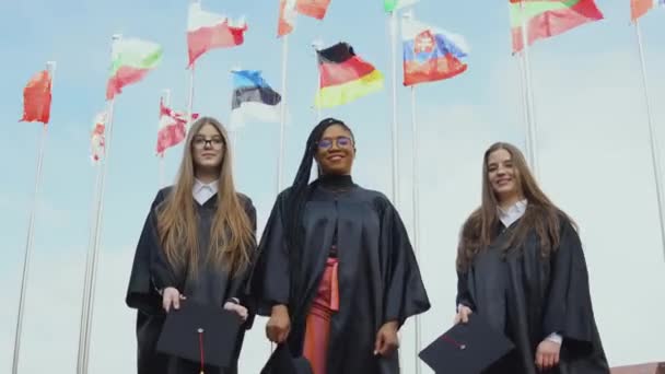 Tre giovani donne di diverse nazionalità lanciano gioiosamente cappelli contro bandiere di diversi paesi che sventolano nel vento sullo sfondo. Vista esterna dal basso — Video Stock