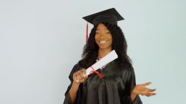 Молодая афроамериканка в черном платье и шляпе мастеров стоит прямо и пожимает диплом выпускника в руке. Она смотрит в камеру. Портрет половины высоты — стоковое видео