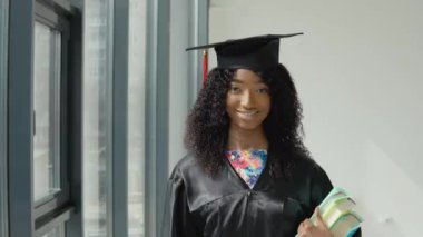 Genç Afrikalı Amerikalı kadın üniversite mezunu bir elinde ders kitaplarıyla duruyor ve büyük parmağıyla siyah çerçeveli modern bir pencerenin yanında iyi bir hareket sergiliyor..