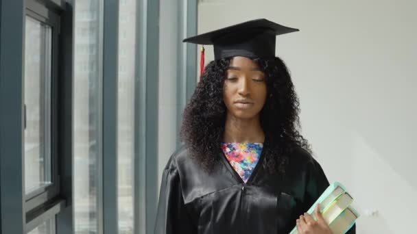 Młoda Afroamerykanka absolwentka uniwersytetu stojąca z podręcznikami w ręku przy dużym nowoczesnym oknie z czarnymi ramkami. — Wideo stockowe