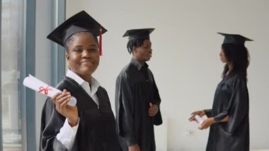 Genç bir Afro-Amerikalı kadın ön planda duruyor ve elinde diplomasıyla kameraya bakıyor. Genç Afro-Amerikan erkek ve kadın arka planda birbirleriyle iletişim kuruyorlar..