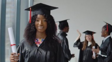 Genç bir Afro-Amerikalı kadın ön planda duruyor ve elinde diplomasıyla kameraya bakıyor. Genç Afrikalı erkek ve kadın ve beyaz bir kadın arkada iletişim kuruyor..