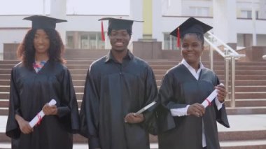 Üç Afro-Amerikan üniversite mezunu dışarıda merdivenlerin karşısında duruyor. Öğrenciler üstat üniforması giymişler.