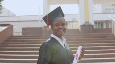 Genç Afro-Amerikan bayan mezun elinde kitaplarla ve diplomasıyla kameranın önünde duruyor. Öğrenci siyah bir bornoz ve kare bir şapka giymiş ve dışarıda duruyor..