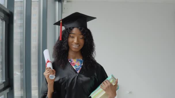 Joven mujer afroamericana graduada de pie frente a la cámara con un diploma y libros en sus manos. El estudiante lleva una túnica negra y un sombrero de maestro cuadrado.. — Vídeo de stock