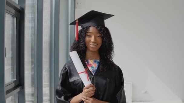 Joven mujer afroamericana graduada de pie frente a la cámara con un diploma en sus manos. El estudiante lleva una túnica negra y un sombrero de maestro cuadrado.. — Vídeo de stock