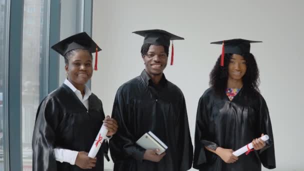 Τρεις Αφροαμερικανοί απόφοιτοι πανεπιστημίου με πτυχία και βιβλία στα χέρια στέκονται δίπλα σε ένα παράθυρο με γκρι πλαίσιο. Οι μαθητές είναι ντυμένοι με γιορτινή ρόμπα και καπέλο. — Αρχείο Βίντεο