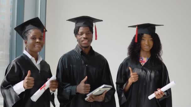 2人の若いアフリカ系アメリカ人女性と外交官とその手に本を持つ1人の若いアメリカ人卒業生が窓のそばに立っている。学生はお祝いの服とマスター帽子を着ています — ストック動画