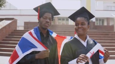 Afrika kökenli Amerikalı erkek ve kadın, siyah cüppeli ve kare şapkalı, omuzlarında İngiliz bayrağı olan öğrencilerle yan yana dururlar.