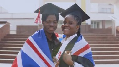 Afrika kökenli Amerikalı erkek ve kadın, siyah cüppeli ve kare şapkalı, omuzlarında İngiliz bayrağı olan öğrencilerle yan yana dururlar.