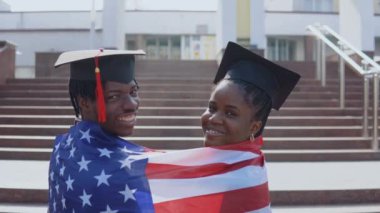 Sırtları kameraya dönük, omuzlarında ABD bayrağı olan mezun öğrencilerin şapkalarıyla Afrika kökenli Amerikalı kadın ve erkek. Kameraya bakıyorum.