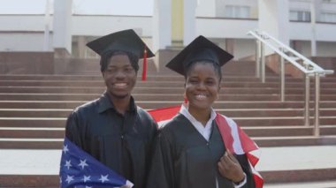 Afro-Amerikalı erkek ve kadın omuz omuza durarak siyah cüppeli ve kare şapkalı öğrencilerin omuzlarında Amerikan bayrağı ile kameraya bakıyorlar.