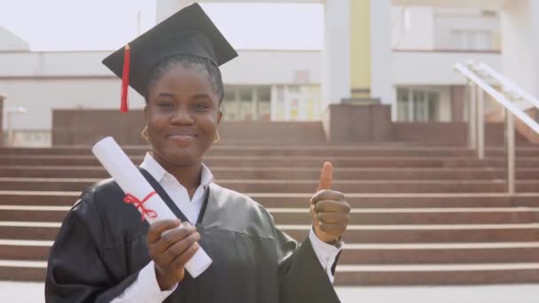 Jovem afro-americana graduada em pé na frente da câmera com um diploma e livros em suas mãos. O estudante está vestido com um manto preto e um chapéu mestres quadrados e fica fora. — Vídeo de Stock